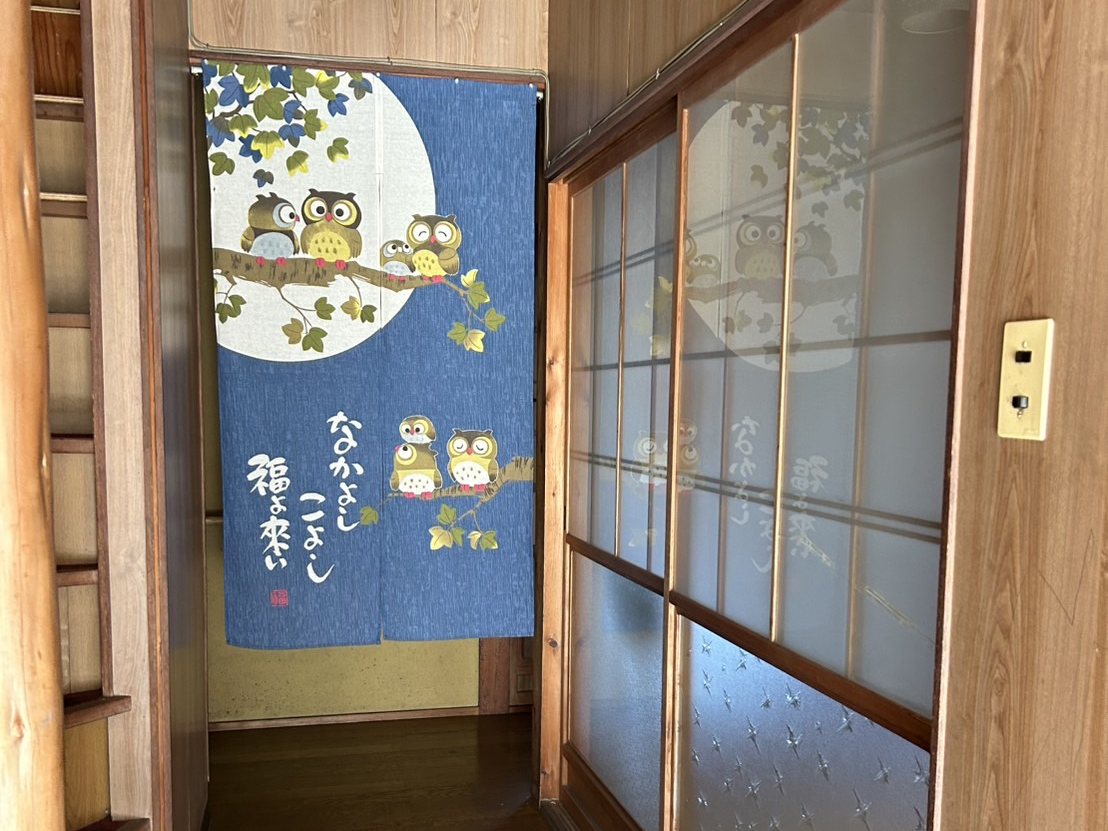 古民家の玄関から続く廊下の写真。左手に階段、正面にふくろう柄の紺色ののれんが掛かっている。